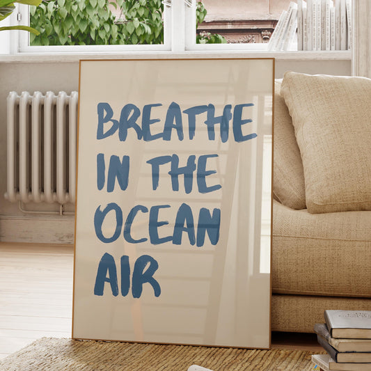 A framed art print leaning against a beige sofa in a bright living room. The print has a cream background with the phrase 'BREATHE IN THE OCEAN AIR' in large blue hand-lettered text.