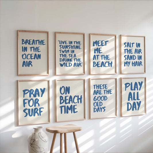 Eight framed wall art prints with blue hand-lettered surf-themed quotes arranged in two rows on a white wall, with a wooden stool and vase below in a sunlit room.