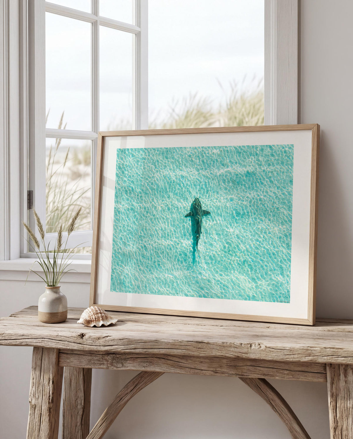 A framed art print depicting a shark swimming in turquoise ocean water, styled on a rustic wooden table next to a small vase with dried grass and a seashell, in front of a window.